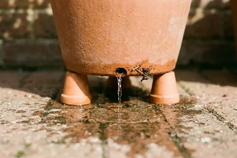 Base of a potted vegetable container resting on pot feet with drainage water flowing from bottom holes onto patio bricks.